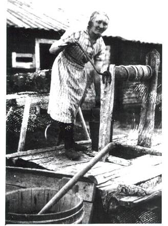 image045.jpg -  Här hinkar Heikan Pekkas och Greta Johannas dotter Ottilia 1912-19xx upp vatten från brunnen i Heikan Pekkas. Bakom syns ladugården. Kan det vara rengöring av trasmattor som är på gång. Några trasmattor syns vid brunnen. Ottilia, yngsta barnet, gifte senare med William Pudas, senare Fällen. De flyttade till Kiruna.