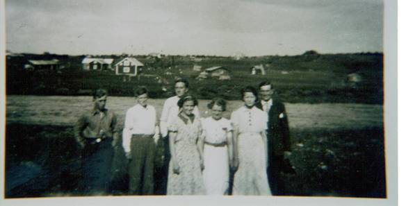 image057.jpg -  Lainioungdomar står på skolgården fotograferade runt 1940 Längst höger William Pudas, senare Vinelund. I mitten, den långa, Manfred Johansson, Flickan i vänster Brita Halldén, Längst vänster Hugo Pudas, sedan Erik Jakobsson, Brita, Ellen Pudas, Pudas, senare Ejdeholm, Bredvid William möjligen Anna Jakobsson Bakom syns Strandelins hus och Kruukas hus på andra sidan älven. Den låg först vid älvstranden, flyttades av Levi Kruuka längre upp som bilden visar. Huset flyttat ännu längre bak när Levis son Elis byggde nytt. Fotograf okänd.