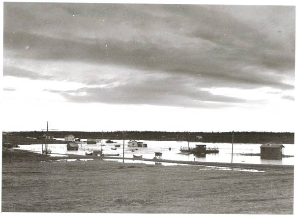 image089.jpg -  Ovanligt högt vattenstånd i Lainioälv augusti 1957 Landsvägen delvis med vatten. Karl Uusitalos sommarhus, till höger, klarar sig, det visste redan TemmingiErkki 1870-talet när han byggde sitt rökpörte precis där Kalle har sin stuga att detta var rätta stället att sätta upp en stuga nära älven. Färjan i sitt sommarläge har ett helt vatten mellan sig och den tillfälliga strandlinjen. Vänster färjan är muraren Helmer Karlssons sommarstuga med vatten in på sig. 2 stycken båtar anslutna vid 3-vägskälet med väg norrut, väg söder och väg mot Vittangihållet. Hit till vägkorsningen kom bäcken från Neitijärvi. Platsen kallades Nikuträsk under 1700-1800 talet. Strax till vänster låg Nikugården som är kvar än men syns inte på bilden. Anordning (mjölkpall) invid vägen, mitt på bilden, där mjölhämtare placerades. Ungefär där bilen finns och till höger står nu hembygdsgården och campingstugorna. Till vänster ses ett hus med gavel och ena långsidan. Det är Sven Aspviks sommarhus. Längre uppåt Johannes Ylipääs sommarhus vid stranden. Mycket ovanligt med denna höga vattennivå i älven. Snön i fjällen smälte bort ovanligt sent. Samuel Eriksson och några av sönerna åkte båt till hans naturängar i Sietki. Alla ladorna hade vatten i sig med det bärgade höet vattenskadat. Det gick åka med båt överallt över ängarna. På andra sidan, östra, syns Millebrands ganska nybyggda hus, till höger ses Karl och Mildas hus flyttat hit från där Millebrands hus nu är. Då 1957 bodde Milda där innan hon bosatte sig i Malmberget. Lorens Vinsa från Vittangi var den som hade hand om skadeomfattningen på skördeskadorna i samband med översvämningarna på ängar. Ängen nedan tillhör Erik och Astrid Uusitalo, De hade kreatur då.