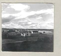 image126.jpg -  Året 1957, augusti. Vita huset närmast Helmer Polströms, vita avlånga huset kapellet i Lainio, innan den en låg byggnad med gräsbevuxet tak är den gamla rian som fortfarande finns kvar, såldes senare av bröderna Isak, Levi och Samuel till hembygdsgården i Jukkas men sattes aldrig upp där. Den gamla kornåkern låg till höger om landsvägen. Det var till rian kornet togs för behandling. Längre bak lång mörk byggnad är Ollagården, ännu längre bak Halldéns hus (med gaveln synlig) Till höger kapellet Valfrid Fällens sommarhus. Längre bort skymtar norra delen av byn. En bit längre ner än bilden visar byggdes ett bevakningstorn under början 1940-talet. Strax höger hitom vägen utanför bilden Erland och Ester Kullebjörk sitt bostadshus. Nyslaget och hässjat hö syns på flera ställen. Till höger Polströms hus låg Lainios första skolbyggnad från 1894. Flyttat 1909 till dess nuvarande plats på skolområdet.