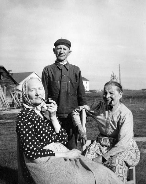 image138.jpg -  Foto Hilma Fredriksson, möjligen 1956. Bild från Lainioboken. Här sitter, i början 1950-talet, Maria Olla (Pirtti Maria) 1888-1956 gift 1907 med Isak Fredriksson 1885-1969, till höger Maria är Hulda Junkka 1891-1978 gift 1921 med Isak Olla 1893-1976 på Ollagården. Isak bror till Maria. Bakom Ollas uthusdel, längre bak bönhuset och vita huset längst bak Erik Jakobssons hus. Båda damerna röker pipa.